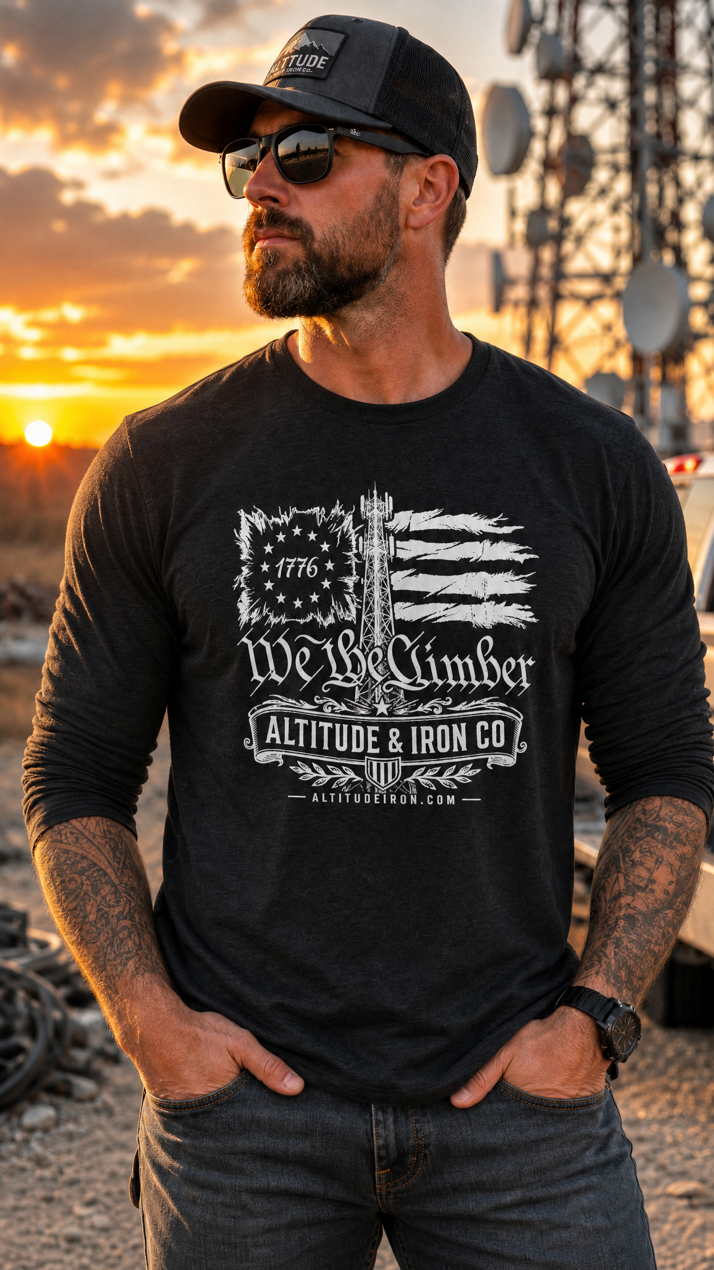 A man wearing a black long-sleeve graphic t-shirt with a distressed American flag and mountain design, featuring the text 'We The Climber' and 'ALTITUDE & IRON CO'. He is also wearing a black trucker hat and sunglasses, standing outdoors at sunset with a communication tower in the background.