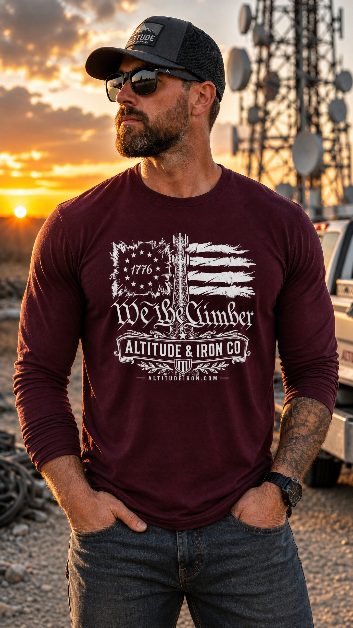 A man wearing a maroon long-sleeve graphic t-shirt with a distressed American flag and 'Altitude & Iron Co' logo, standing outdoors at sunset near a truck and communication towers.