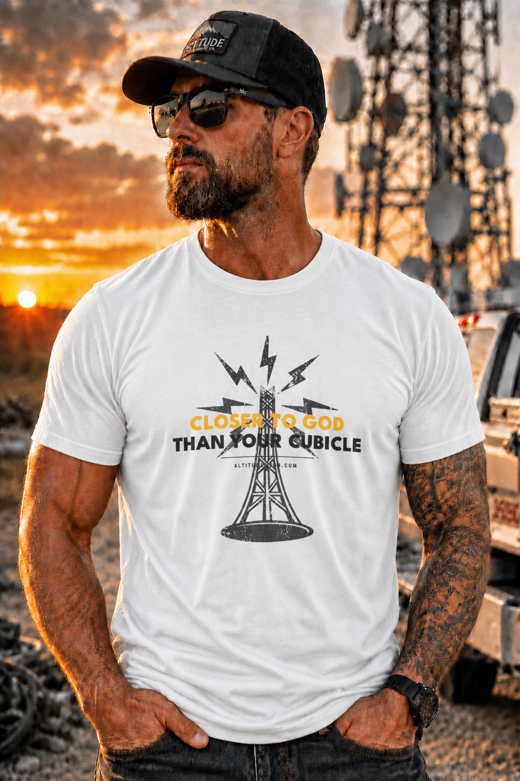 A man wearing a white graphic t-shirt with a black cap and sunglasses, posing outdoors against a sunset background with a cell tower.